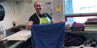 Female volunteer holding wearing a light green apron with text that say "Volunteer" holding a blue town that she is in the middle of folding.