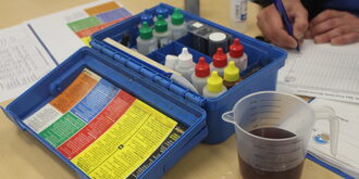 A hand writes on a form in the corner beside a pool water testing kit. 