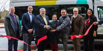 Smiling County, municipal and transportation leaders are cutting a red ribbon stretched across in front of the SPOT Southwest shuttle van These leaders are (left to right): Metro Transportation Policy Director Ted Leybold; Ride Connection CEO Andy Nelson; Washington County Commissioner Jason Snider; Beaverton Councilor Ashley Hartmeier-Prigg; Tigard Mayor, Yi-Kang Hu; Washington County Land Use & Transportation Director Stephen Roberts; Safe Routes to Schools Coordinator, Leah Biado-Luis.
