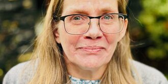 A middle-aged Caucasian woman with blonde hair wearing glasses looking at the camera.