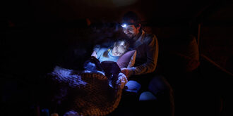 A father and daughter cuddle in the dark using a headlamp to read