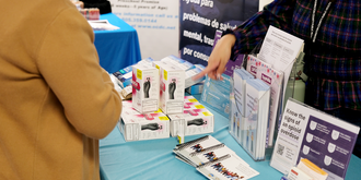 A woman in a brown coat looks over as a person across them points at a table full of materials and resources related to substance use prevention. 