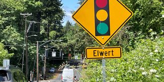 A view down Edy Road looking toward its intersection with Elwert Road. Close by on the right side is a big traffic sign showing that a traffic signal is ahead and below it is a sign saying that Elwert is the cross street ahead.