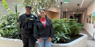 Man in police uniform and woman in street clothes smile at camera in front of plants