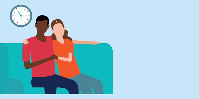 A man and woman sit on the couch holding hands, as a clock hangs behind them. 