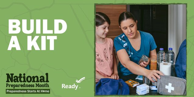 A mother and daughter sit at a kitchen table that is covered in preparedness supplies like a first aid kit, toilet paper, and water bottles