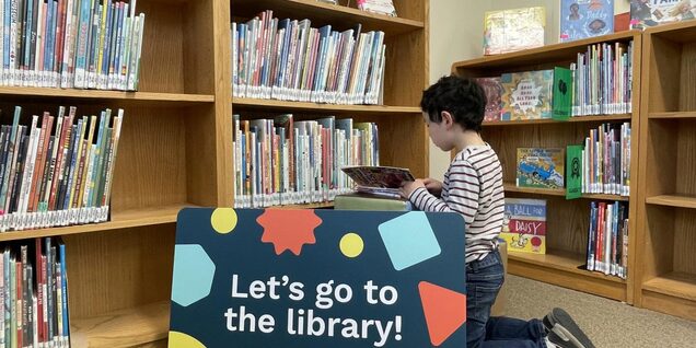 Child kneeling in the library reading next to a sign that reads "Let's go to the library!"