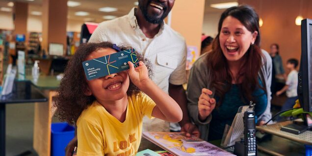 Child with a library card over their eyes with parents smiling behind them