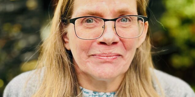 A middle-aged Caucasian woman with blonde hair wearing glasses looking at the camera.