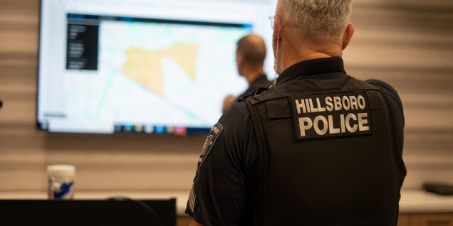 Back of a man wearing a hillsboro police uniform looking at a large screen showing a map