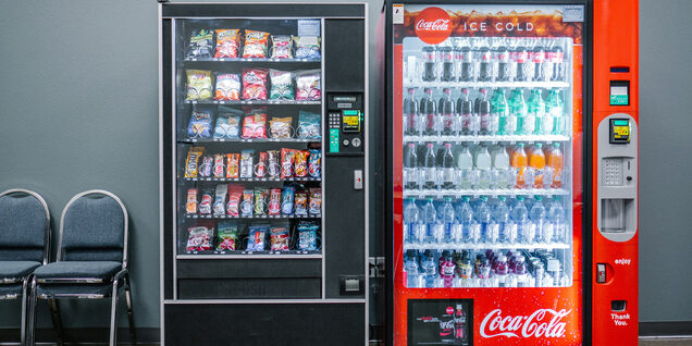 Two vending machines for snacks and sodas sit side by side in a grey waiting area.