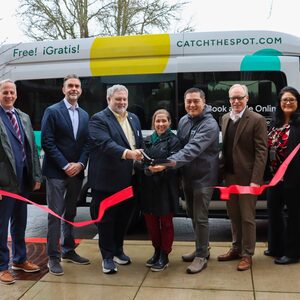 Smiling County, municipal and transportation leaders are cutting a red ribbon stretched across in front of the SPOT Southwest shuttle van These leaders are (left to right): Metro Transportation Policy Director Ted Leybold; Ride Connection CEO Andy Nelson; Washington County Commissioner Jason Snider; Beaverton Councilor Ashley Hartmeier-Prigg; Tigard Mayor, Yi-Kang Hu; Washington County Land Use & Transportation Director Stephen Roberts; Safe Routes to Schools Coordinator, Leah Biado-Luis.