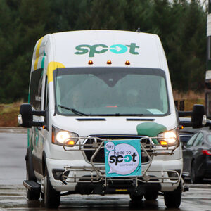 The SPOT microtransit passenger van style shuttle, a white van with yellow, green and aqua colored polka-dots driving toward you with its headlights on. A sign on the front reads "Say HI to SPOT!"