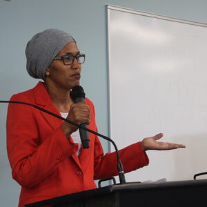Commissioner Nafisa Fai takes the stage, wearing a red blazer and a gray head cover while holding a microphone in her hand. 