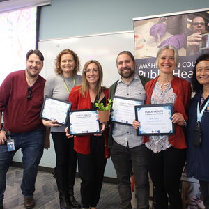 Six people stand before the camera for a photo, with four of them holding a plaque reward they've received. 