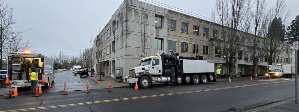 Large repair trucks are parked along the Washington County Public Services Building along West Main Street in Hillsboro, Oregon. Public works personnel are walking along the roadway and sidewalk as they engage in emergency plumbing operations. Reflective traffic cones line part of the roadway.