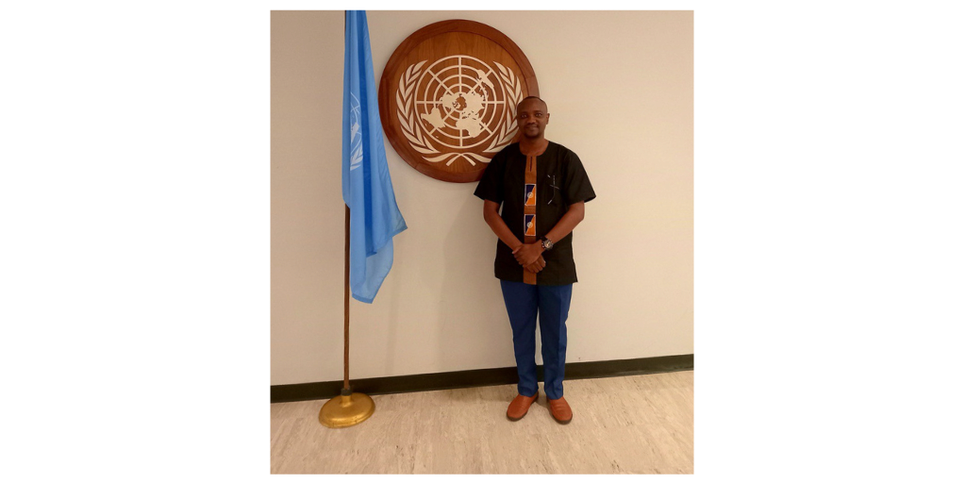A man wearing a black shirt stands in front of a mounted logo on the wall and a related flag of the World Health Organization (WHO) 