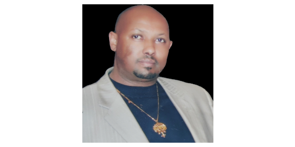 A man wearing a gray blazer over a black shirt and yellow necklace. 
